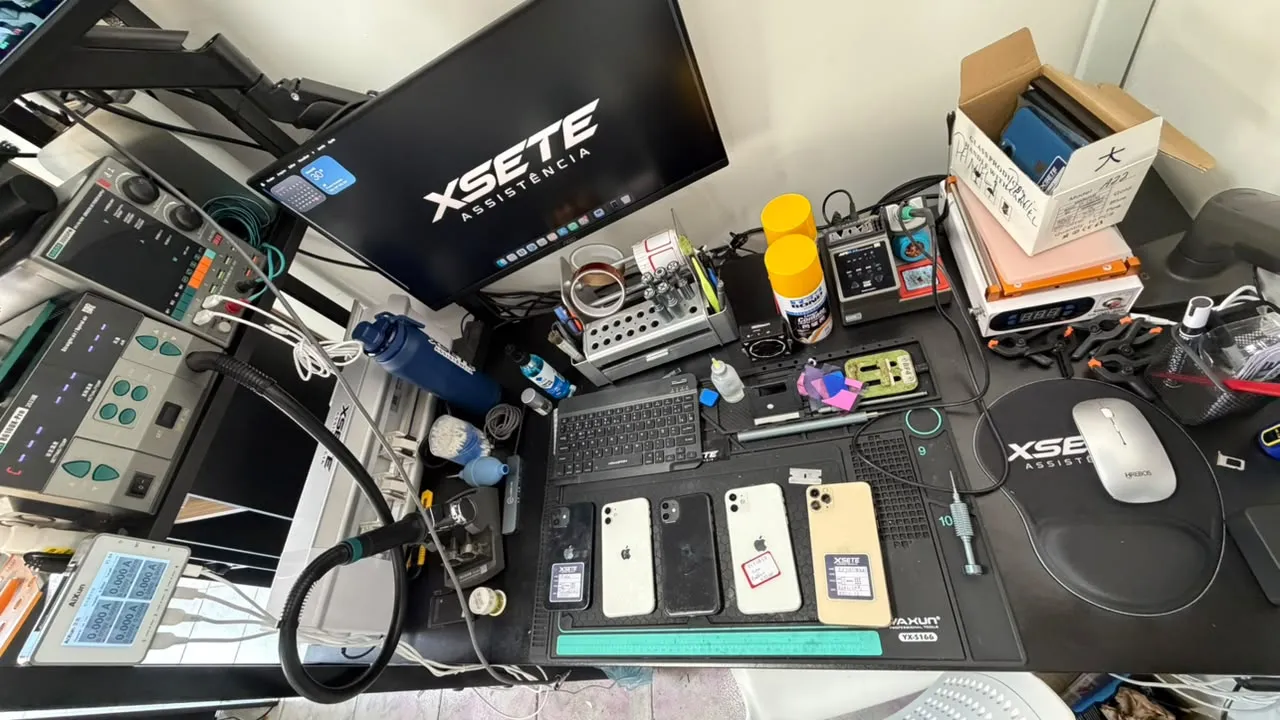 Bancada de reparo da XSETE vista de cima, com cinco iPhones identificados, ferramentas e monitor com a marca XSETE