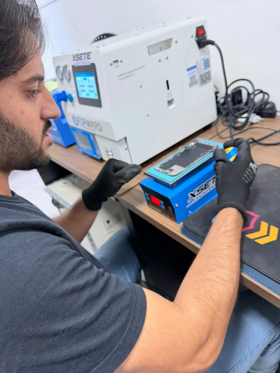 Técnico da XSETE de luvas trabalhando em uma tela de celular sobre a bancada, ao lado de equipamentos com a marca XSETE