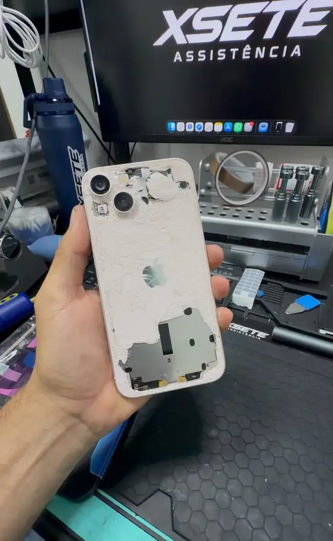 Mão segurando um iPhone com o vidro traseiro todo trincado e partes soltas, na bancada da XSETE